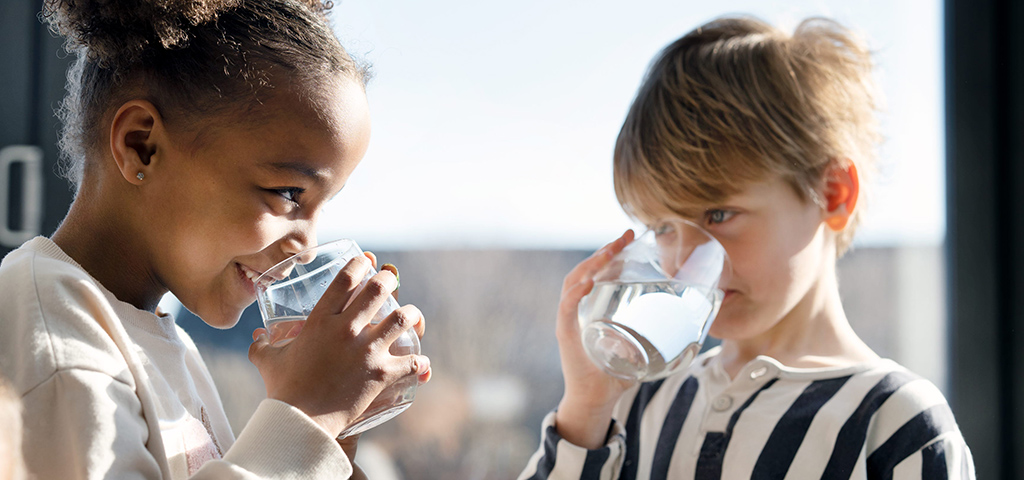 Två barn står och tittar leende på varandra samtidigt som de dricker vatten från varsitt glas.