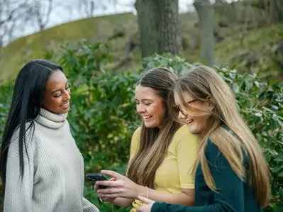 Tre elever skrattar medan de tittar på något på en mobiltelefon utomhus.